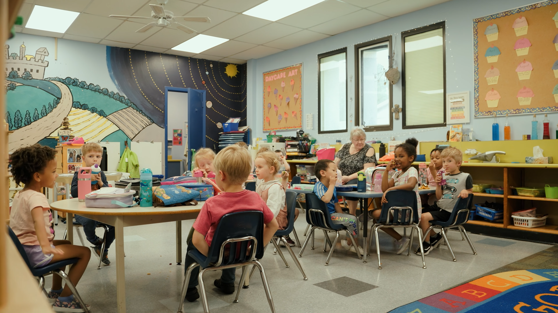 Children sitting at tables in daycare with one teacher.
